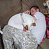 dog, man, sleeping, couch, blanket, pet, companion, indoors, cozy, nap, resting, white_dog, brown_ears, collar, cuddle, living_room, furniture, snacks, casual_clothing, portrait