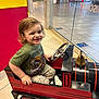 Owen joined the competition — help win amazing prizes! toddler, child, train_ride, winking, smiling, indoor, mall, toy, red, green_shirt, playful, floor_tiles, glass_door, shopping_mall, steering_wheel, bell, short_hair, casual_clothing, fun, cute