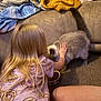 child, girl, blonde_hair, cat, pet, couch, blanket, indoor, carpet, food_bowl, animal, furniture, long_hair, hand, feeding, domestic_animal, cozy, home, interaction, friendly