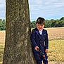 boy, casual_pose, child, daytime, field, formal_wear, grass, nature, outdoor, playful, portrait, shoes, sky, smile, suit, sunny, tongue_out, toy_gun, tree, young