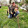 animal, black_dog, cap, casual, dog, flowers, glasses, grass, house, kneeling, leash, nature, outdoor, person, pet, pink_harness, small_dog, spring, tattoo, tree