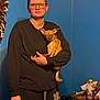 Phoebe joined the competition — help win amazing prizes! person, dog, glasses, blue_wall, black_clothing, short_hair, small_dog, indoor, table, bottle, furniture, casual, clutter, hand, ears, pet, holding, standing, expressionless, room