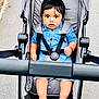 baby, child, stroller, blue_outfit, seatbelt, barefoot, outdoor, pavement, curious, young_child, cute, infant, walking_path, black_hair, sitting, safety_harness, summer_clothing, daylight, portrait, person