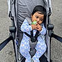baby, child, stroller, pacifier, onesie, blue, gray, outdoor, pavement, infant, seatbelt, cute, young_child, fence, casual, resting, clothing, footwear, sitting, baby_girl