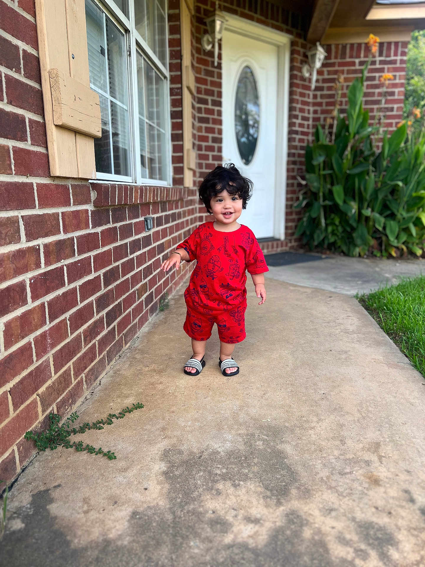 Jesús is registered to the contest to win money with this photo: toddler, child, red_clothing, sandals, curly_hair, smiling, outdoor, brick_wall, house, door, concrete_path, plants, greenery, sidewalk, portrait, person, happy, daylight, front_yard, casual
