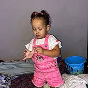 Zelyiana is registered to the contest to win money with this photo: toddler, child, pink_overalls, white_tshirt, barefoot, bed, blankets, bucket, indoor, curly_hair, hair_ties, focused, playing, casual_clothing, young_child, home, floor, furniture, portrait, person
