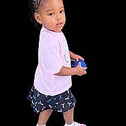 Harminey joined the competition — help win amazing prizes! toddler, child, girl, braided_hair, pink_ribbons, pink_shirt, patterned_skirt, white_socks, pink_shoes, toy, standing, curious, portrait, isolated_background, black_background, casual_clothing, young_child, cute, looking_back, full_body