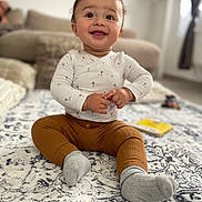 Amir a rejoint le concours — aidez-le/la à gagner de superbes lots ! baby, child, smiling, sitting, indoor, carpet, rug, clothing, pants, shirt, socks, face, hands, person, home, couch, blurred_background, window, curtains, toy