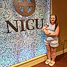 Wrenley is registered to the contest to win money with this photo: adult, baby, mother, infant, parent, portrait, standing, holding, blanket, hospital, nicu, sign, indoor, wall, colorful, headband, smiling, clothing, wood_floor, family