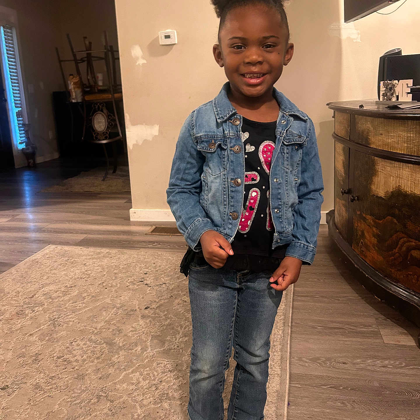 Aesia is registered to the contest to win money with this photo: black_shirt, cabinet, casual_clothing, child, denim_jacket, girl, hair_up, happy, home, indoor, jeans, natural_light, pattern, portrait, room, rug, smiling, sneakers, standing, wooden_floor