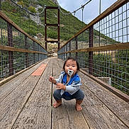 David is registered to the contest to win money with this photo: toddler, child, pacifier, bridge, wooden_bridge, mountain, outdoor, nature, baby, fence, cable, cloudy_sky, suspension_bridge, curious, squatting, casual_clothing, barefoot, greenery, hill, daytime