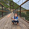 toddler, child, pacifier, bridge, wooden_bridge, mountain, outdoor, nature, baby, fence, cable, cloudy_sky, suspension_bridge, curious, squatting, casual_clothing, barefoot, greenery, hill, daytime