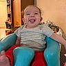 baby, smiling, tongue_out, sitting, baby_seat, gray_shirt, red_pants, indoor, furniture, cabinet, baby_bottle, happy, cute, child, person, home, table, foot, head, room