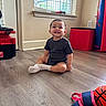 Adriel is registered to the contest to win money with this photo: toddler, child, smiling, sitting, indoors, portrait, playroom, floor, wood_floor, window, blinds, sunlight, black_shirt, white_socks, toy, play_equipment, toolbox, red, blue, happy