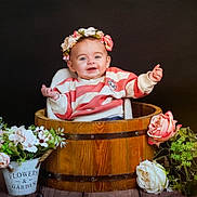 Marie participe au concours pour gagner de l'argent avec cette photo : baby, child, cute, decorative, flower_crown, flowers, greenery, happy, indoor, pink_flowers, plant, portrait, pot, sitting, smiling, striped_sweater, white_flowers, wooden_bucket, wooden_surface, young_child