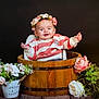 baby, child, cute, decorative, flower_crown, flowers, greenery, happy, indoor, pink_flowers, plant, portrait, pot, sitting, smiling, striped_sweater, white_flowers, wooden_bucket, wooden_surface, young_child