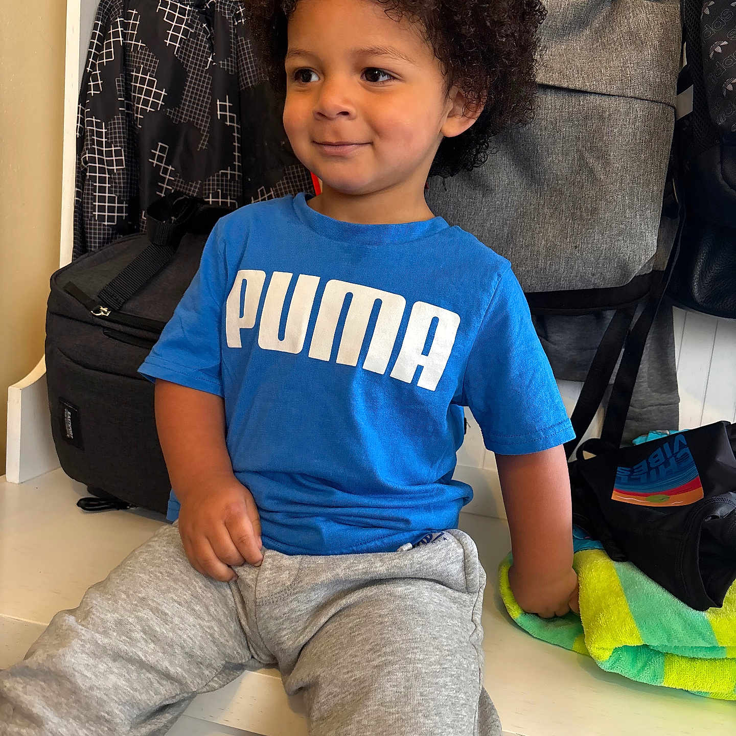Karter Deqwan Dixon joined the competition — help win amazing prizes! bags, bench, blue_tshirt, casual, child, clothing, curly_hair, daylight, grey_sweatpants, happy, home, indoor, person, portrait, puma_logo, relaxed, seated, smiling, towel, young_child