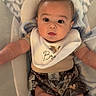 arms, baby, baby_seat, bib, blanket, bottle, camo_shorts, child, clothing, cute, expression, face, floor_tiles, head, indoor, infant, legs, person, sitting, young