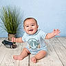 baby, bare_feet, blue_background, child, decorative_duck, face, happy, houseplant, indoor, infant, onesie, outfit, photography, portrait, sitting, smiling, studio, toy, waving, wooden_floor