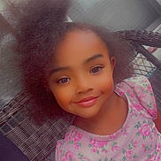 Alianna is registered to the contest to win money with this photo: child, girl, smiling, portrait, selfie, sitting, woven_chair, floral_shirt, pink_clothing, curly_hair, hair_puffs, brown_eyes, indoors, window, chair, close_up, happy, emojis, casual, photograph
