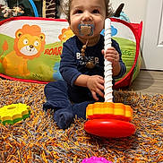 Eden participe au concours pour gagner de l'argent avec cette photo : carpet, child, colorful_rug, curly_hair, floor, happy, indoors, lion_graphic, nursery, onesie, pacifier, pacifier_clip, play_tent, playroom, plush_giraffe, sitting, smile, stacking_toy, toddler, toy