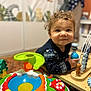 abacus, baby, bead_maze, child, close_up, colorful_toy, curly_hair, hand, happy, indoor, looking_at_camera, playmat, playroom, portrait, shallow_depth_of_field, smile, sweater, toddler, toy, wooden_table