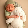 baby, newborn, sleeping, plush_toy, bunny, cuddle, knit_cap, soft_texture, green_clothing, infant, child, cute, peaceful, portrait, indoors, warm_colors, cozy, hands, face, toddler