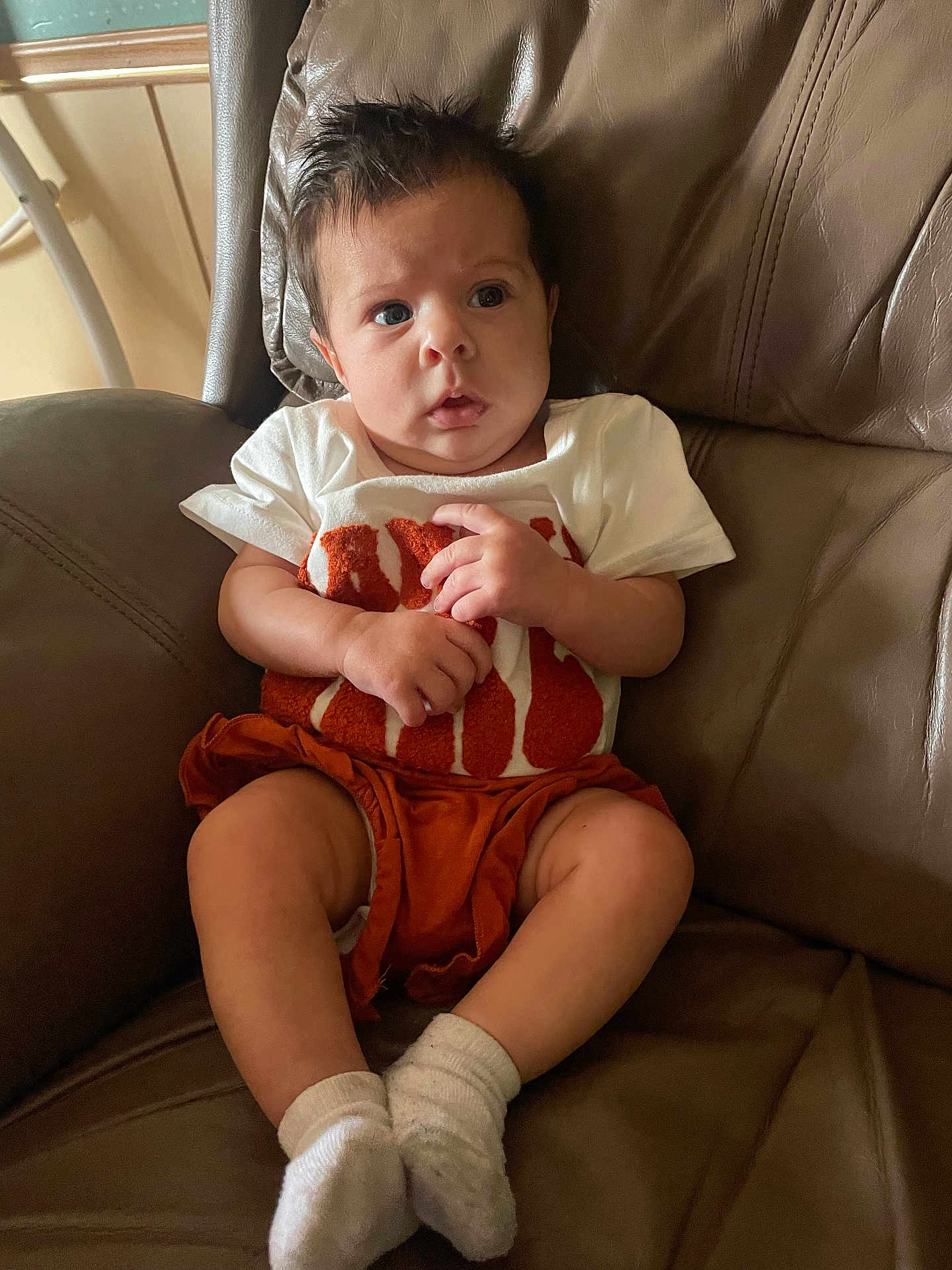 Kehlani joined the competition — help win amazing prizes! baby, child, infant, sitting, chair, leather, white_shirt, orange_shorts, socks, indoor, curious, portrait, toddler, hands, legs, face, hair, casual_clothing, home, furniture