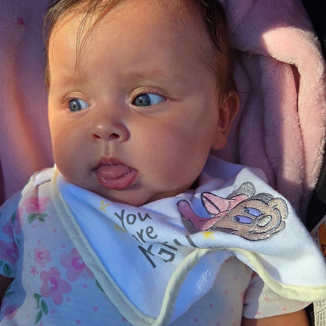 Mia Charlotte is registered to the contest to win money with this photo: baby, bib, blue_eyes, cartoon_character, child, closeup, cute, expression, face, floral_outfit, indoors, infant, newborn, person, pink_blanket, skin, sleepwear, soft_light, tongue_out, young_child