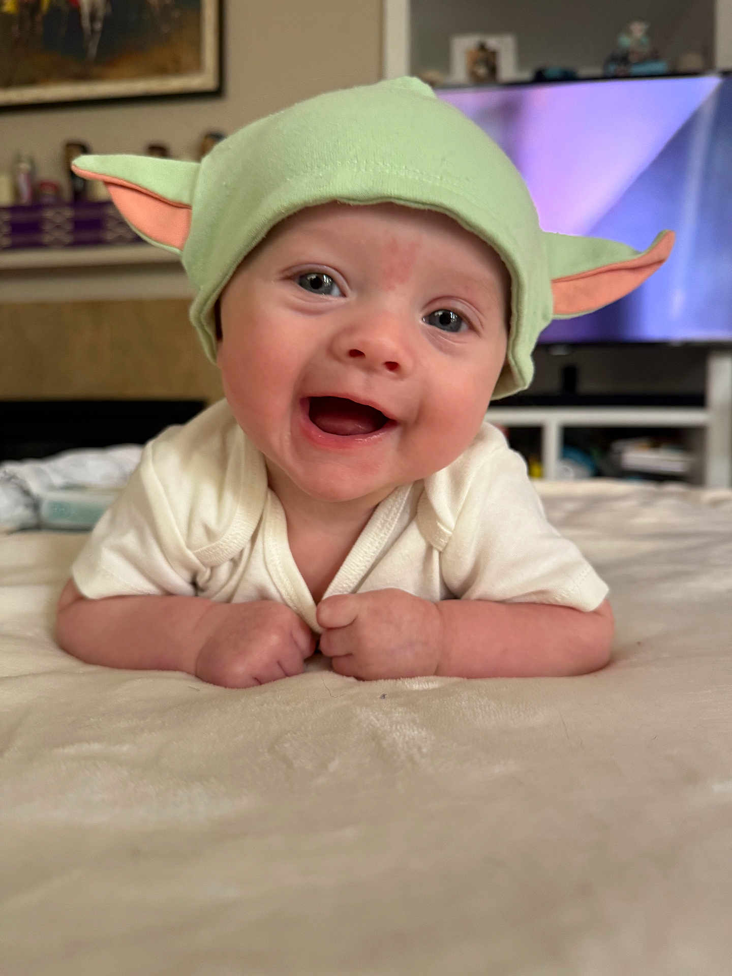 August is registered to the contest to win money with this photo: baby, infant, smiling, green_hat, yoda_hat, onesie, hands, forearms, bed, blanket, closeup, portrait, cute, happy, eyes, living_room, television, furniture, soft_texture, cheerful