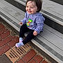 Adrianna is registered to the contest to win money with this photo: toddler, child, smiling, hoodie, colorful, dog_graphic, black_pants, socks, sitting, wooden_steps, brick_pavement, memorial_plaques, outdoor, happy, face, person, casual_clothing, daylight, urban, cute