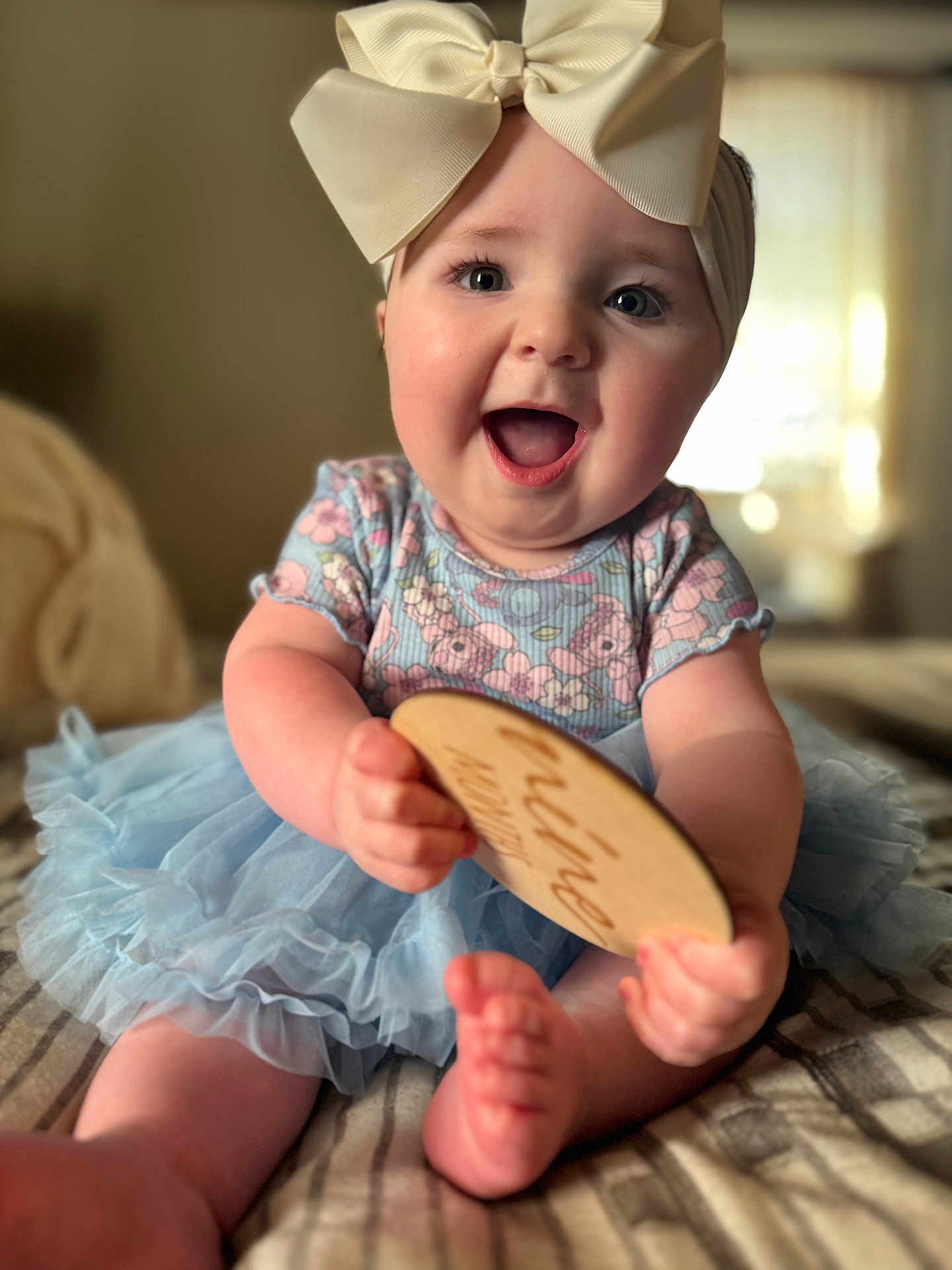 Adrianna is registered to the contest to win money with this photo: baby, child, infant, bow, headband, smiling, happy, blue_dress, floral_pattern, tutu, sitting, barefoot, plaque, wooden_sign, indoors, bed, blanket, portrait, cute, joyful