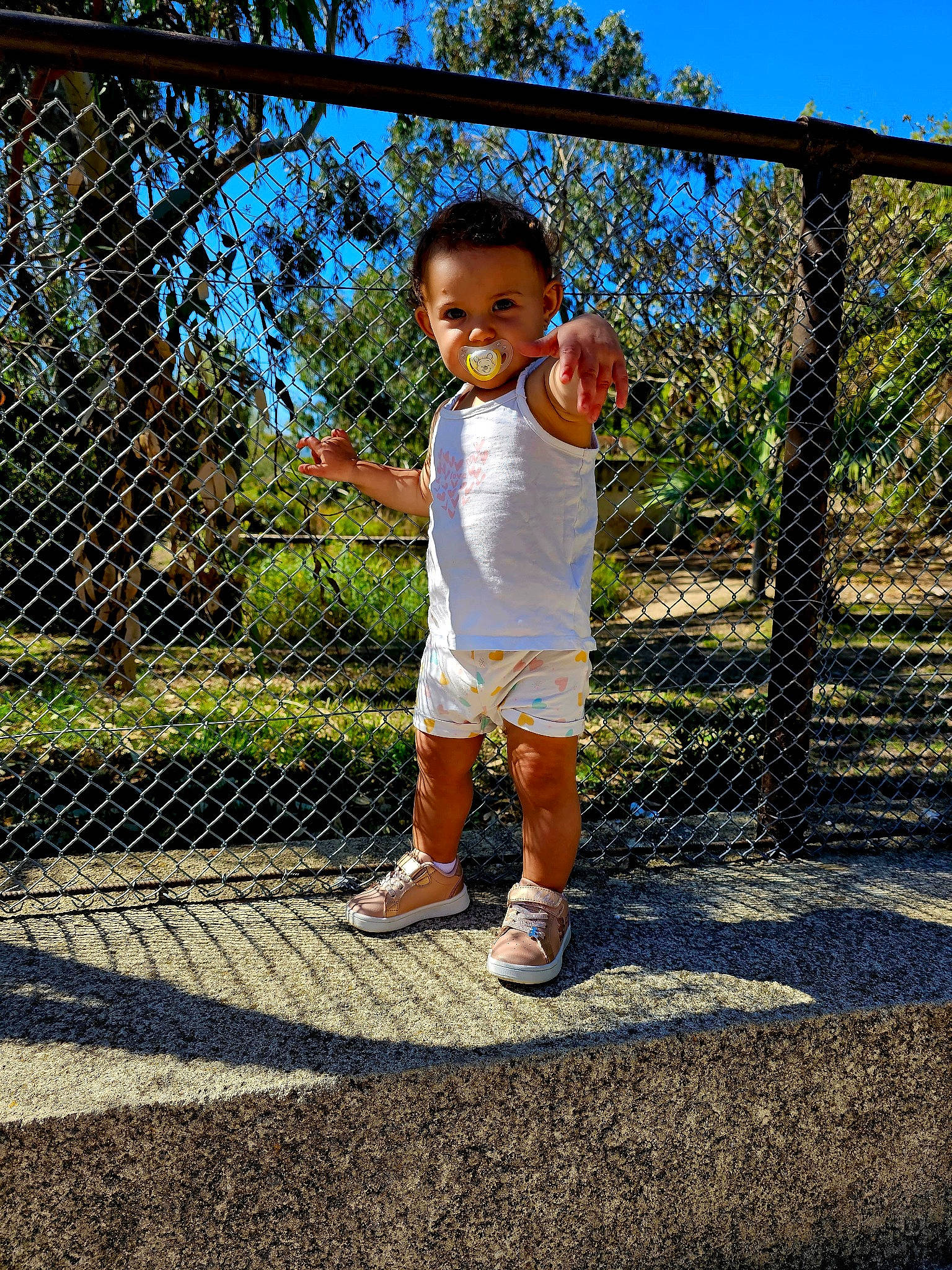 Ruby a rejoint le concours — aidez-le/la à gagner de superbes lots ! chain_link_fencing, fence, fun, grass, happy, landscape, leisure, mesh, nature, people_in_nature, person, plant, public_space, shorts, sky, standing, sunlight, toddler, tree, wire_fencing
