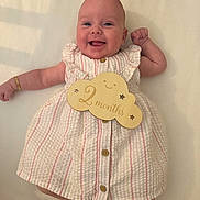 Cora is registered to the contest to win money with this photo: baby, bracelet, button, celebration, child, cloud, cute, dress, happy, infant, lying_down, newborn, person, pink_stripes, portrait, skin, smile, two_months, white_background, wooden_sign