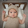 baby, infant, knitted_hat, knitted_outfit, bear_ears, wooden_bed, pillow, lying_down, cute, wide_eyes, raised_arms, indoors, portrait, child, adorable, soft_lighting, neutral_colors, cozy, sleepwear, baby_fashion