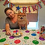 woman, dog, birthday, party_hat, table, cake, birthday_card, balloons, banner, celebration, indoor, smiling, decorations, party, pet, happy, person, festive, colorful, event