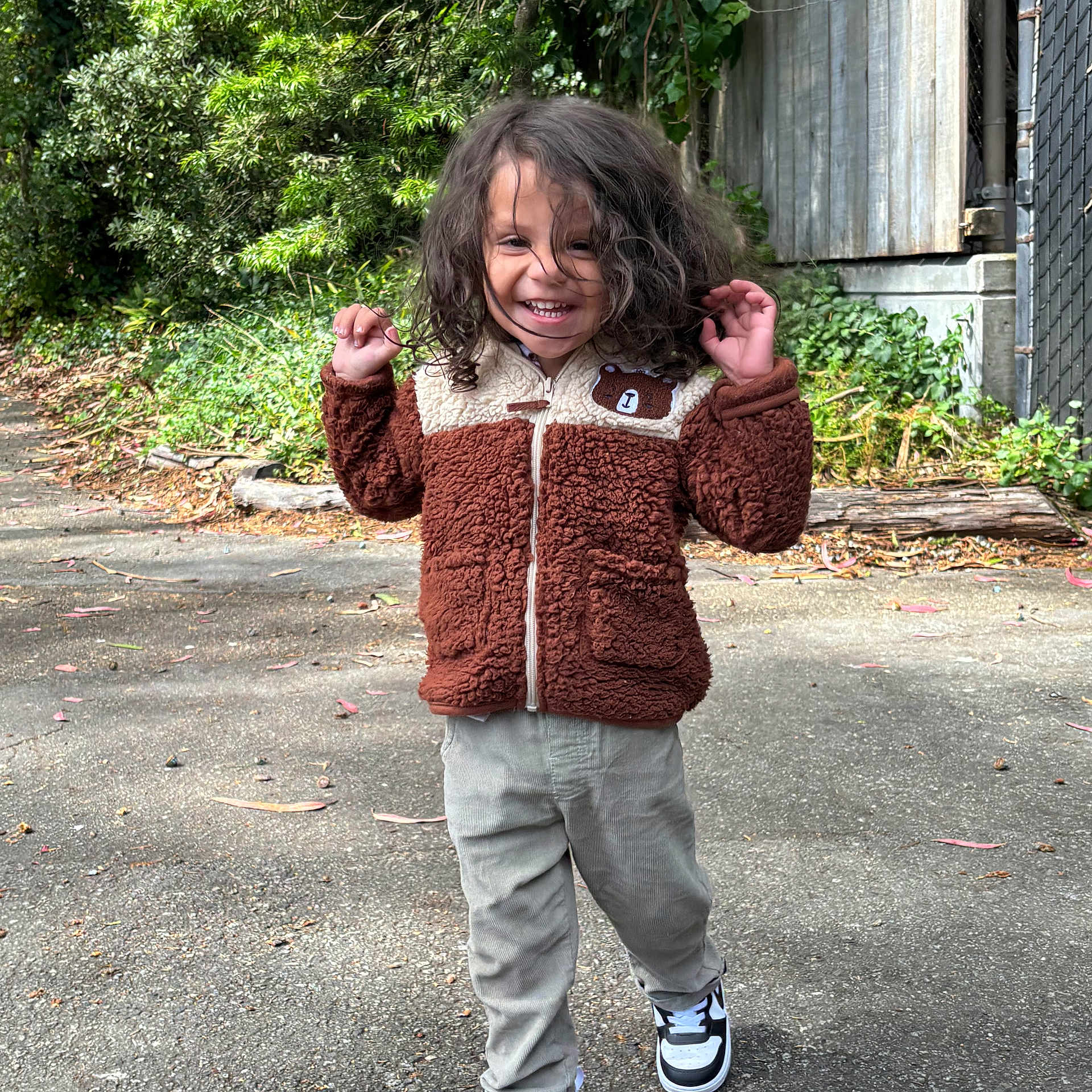 Art Iv is registered to the contest to win money with this photo: child, toddler, curly_hair, smiling, jacket, pants, sneakers, outdoor, path, greenery, fence, wood, sunlight, walking, happy, person, nature, casual_clothing, daytime, cute