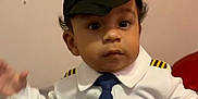 Kamaz is registered to the contest to win money with this photo: baby, toddler, child, pilot_costume, hat, tie, uniform, portrait, face, eyes, hands, sitting, indoor, bed, blanket, pillow, medal, wall, cute, curly_hair
