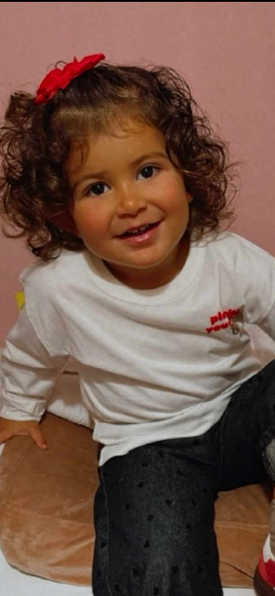 Assia a rejoint le concours — aidez-le/la à gagner de superbes lots ! toddler, child, smiling, curly_hair, red_bow, white_shirt, dark_pants, cushion, indoor, portrait, happy, cute, person, sitting, young_child, casual_clothing, face, skin, head, eyebrows
