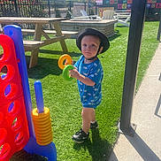 Carmello is registered to the contest to win money with this photo: toddler, child, outdoor, grass, toy, connect_four, picnic_table, hat, blue_onesie, shoes, sunlight, playground, fence, plastic_toy, lawn_chair, sidewalk, shadow, daytime, greenery, recreation