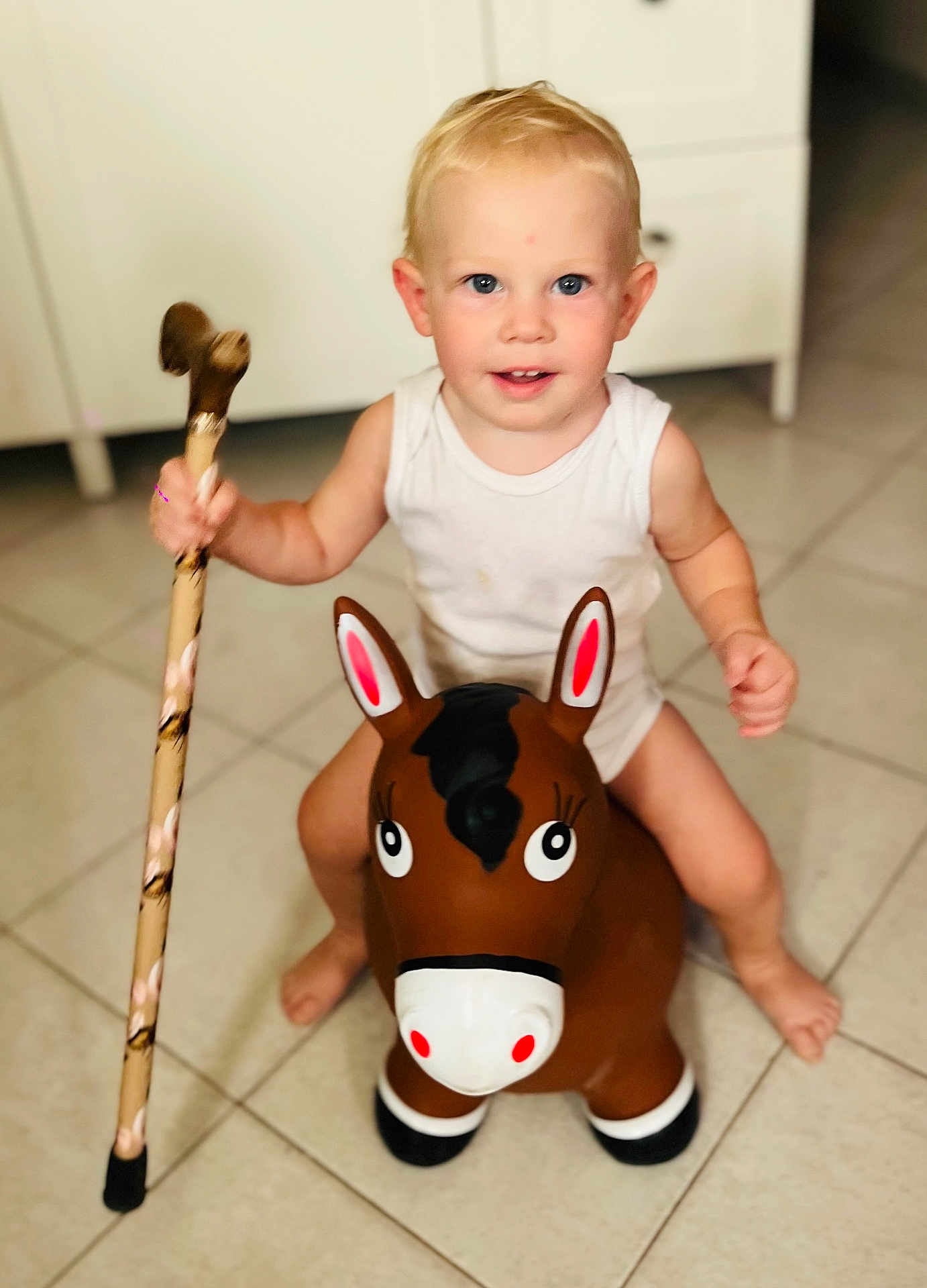 Tom participe au concours pour gagner de l'argent avec cette photo : toddler, child, indoor, toy, inflatable_horse, walking_stick, white_tank_top, floor_tiles, kitchen, barefoot, blond_hair, blue_eyes, smiling, play, fun, cute, person, baby, young_child, home