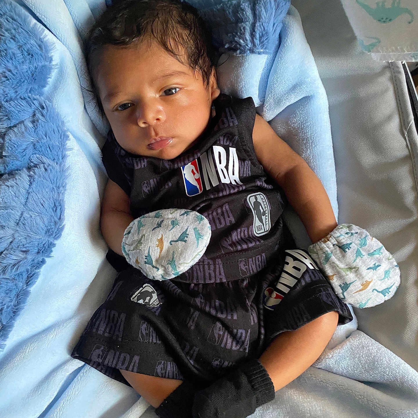 Kassiah is registered to the contest to win money with this photo: baby, baby_clothes, black_socks, blanket, blue_blanket, clothing, cute, indoors, infant, laying_down, mittens, nba_logo, newborn, person, portrait, relaxed, small_hands, soft_texture, sportswear, young_child