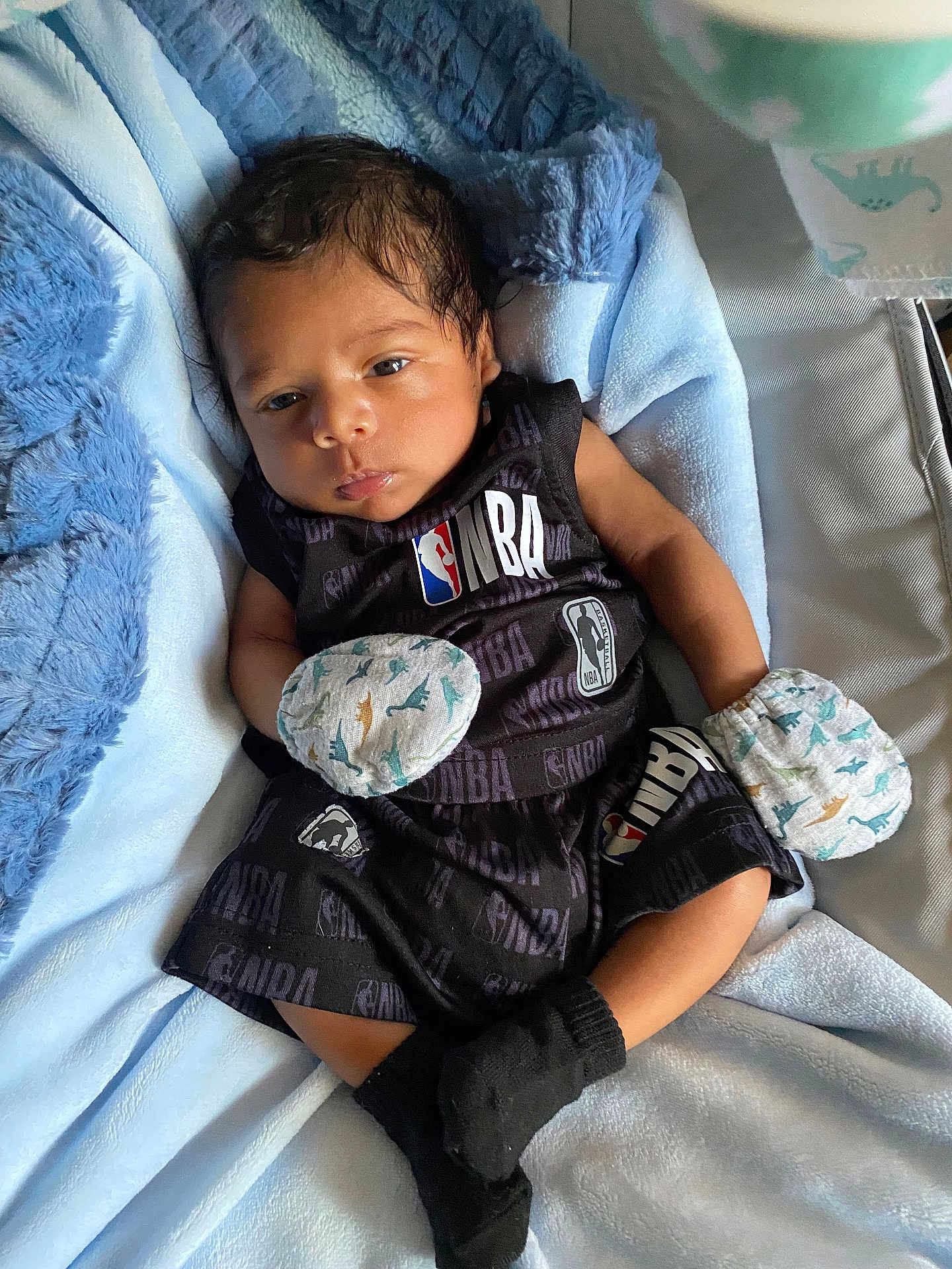 Kassiah is registered to the contest to win money with this photo: baby, baby_clothes, black_socks, blanket, blue_blanket, clothing, cute, indoors, infant, laying_down, mittens, nba_logo, newborn, person, portrait, relaxed, small_hands, soft_texture, sportswear, young_child