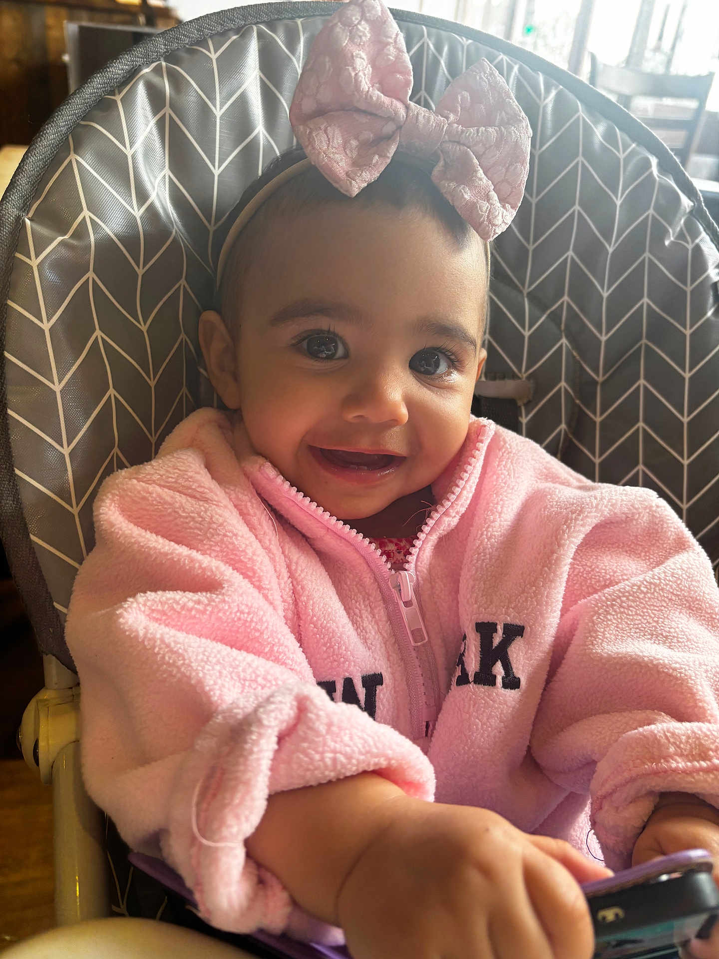 Elula is registered to the contest to win money with this photo: baby, child, high_chair, pink_jacket, bow_headband, smiling, indoor, toy, face, eyes, hand, clothing, person, cute, happy, portrait, infant, seat, soft_texture, natural_light