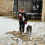 boy, child, dog, puppy, snow, snowball, winter_clothing, gloves, trampoline, stone_wall, outdoor, playful, curious, footwear, pavement, pet, cold_weather, young, fun, activity