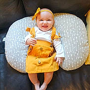 Aurora is registered to the contest to win money with this photo: baby, child, infant, smiling, happy, portrait, indoor, seated, cushion, pillow, mustard_outfit, headband, bow, overalls, white_shirt, leather_couch, furniture, photography, adorable, centered