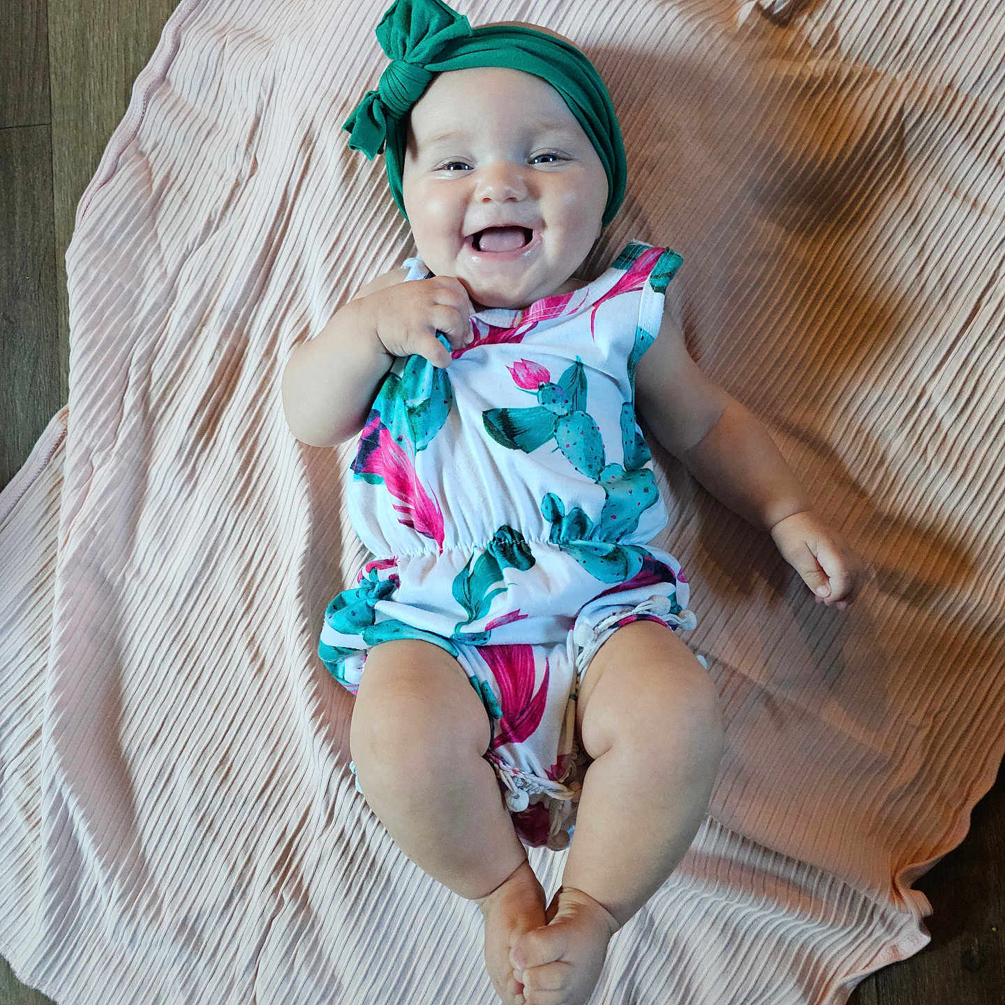 Aurora is registered to the contest to win money with this photo: baby, child, infant, smiling, portrait, lying_down, blanket, pink_blanket, teal_headband, bow, floral_outfit, colorful_clothing, happy, face, hands, feet, indoor, wooden_floor, overhead_view, photography