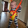 Reece is registered to the contest to win money with this photo: baby, child, striped_clothing, socks, baby_jumper, yellow, orange, lion_toy, hallway, wood_floor, door, indoor, smiling, walking, safety_harness, playful, cute, infant, person, young_child