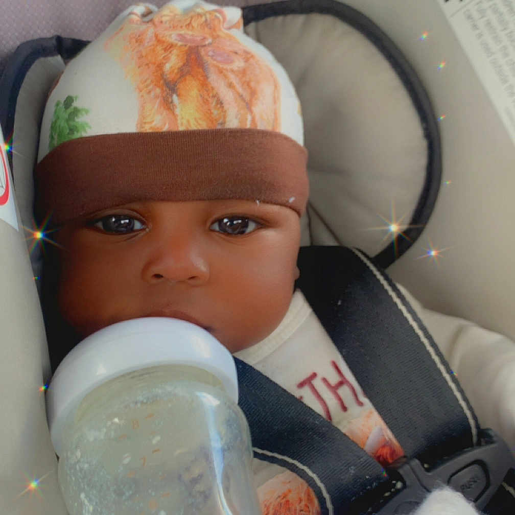 Kamari is registered to the contest to win money with this photo: baby, car_seat, bottle, hat, mittens, child, face, seatbelt, infant, drink, cozy, cute, indoor, portrait, sitting, clothing, person, sparkle_effect, closeup, warm