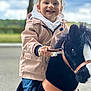 child, smiling, toy_horse, plush_toy, outdoor, jacket, hoodie, jeans, happy, play, cute, young_child, portrait, hands, animal_toy, nature, greenery, daylight, person, fun